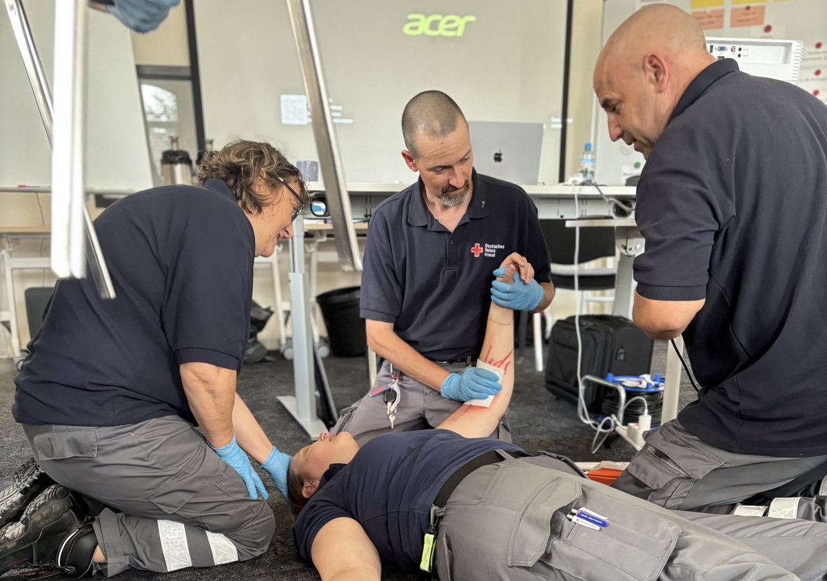 Zwei Sanitätshelfer und eine Sanitätshelferin knien um eine weitere Sanitätshelferin und drücken einen Verband auf eine geschminkte Wunde an ihrem Arm.