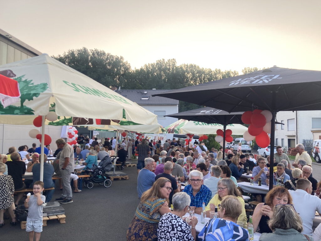 Zahlreiche Gäste auf dem Sommerfest des DRK. Sonnenschirme sind mit weiß-roten Luftballons geschmückt.