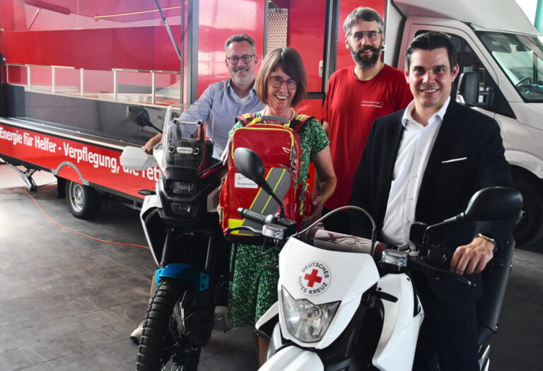 Das Foto zeigt von rechts Marco Laut, Stiftungsvorstand der Sparkasse Odenwaldkreis, DRK-Jugendkoordinator Adrian Knecht sowie mit Sanitätsrucksack Sachbearbeiterin Nicole Kelbert-Gerbig und DRK-Vorstand Frank Sauer. Marco Laut und Frank Sauer sitzen auf DRK-Motorrädern, im Hintergrund ist der rote Küchencontainer zu sehen.