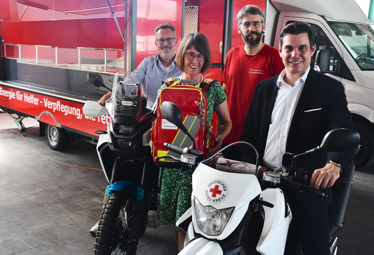 Das Foto zeigt von rechts Marco Laut, Stiftungsvorstand der Sparkasse Odenwaldkreis, DRK-Jugendkoordinator Adrian Knecht sowie mit Sanitätsrucksack Sachbearbeiterin Nicole Kelbert-Gerbig und DRK-Vorstand Frank Sauer. Marco Laut und Frank Sauer sitzen auf DRK-Motorrädern, im Hintergrund ist der rote Küchencontainer zu sehen.