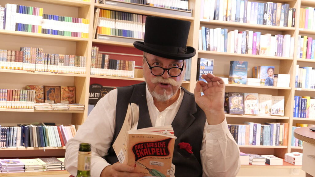 Der Autor im weißen Hemd mit schwarzem Zylinder und schwarzer Weste hält sein Buch vor sich und liest daraus.