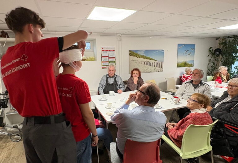 Schulsanitäter führen vor, wie ein Druckverband angelegt wird. Mehrere Leuten schauen interessiert zu.